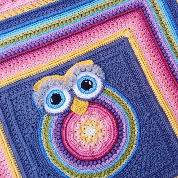 Owlie 2.0 crochet kit A colourful crocheted owl square with a central owl motif in pink, purple, yellow, and blue, set against a dark blue background and bordered with bright yellow, pink, purple, and blue rounds.