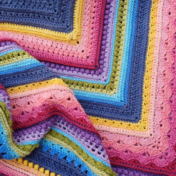 An intricately crocheted square blanket featuring layered rows of varied stitch textures in vibrant colours—blue, pink, purple, yellow, green, and red—displayed flat to show the pattern and craftsmanship