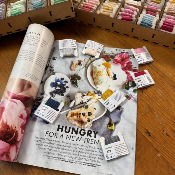 An open magazine spread used as a colour reference, with embroidery thread cards arranged across the page, and wooden storage boxes filled with hand-dyed embroidery threads visible in the background.