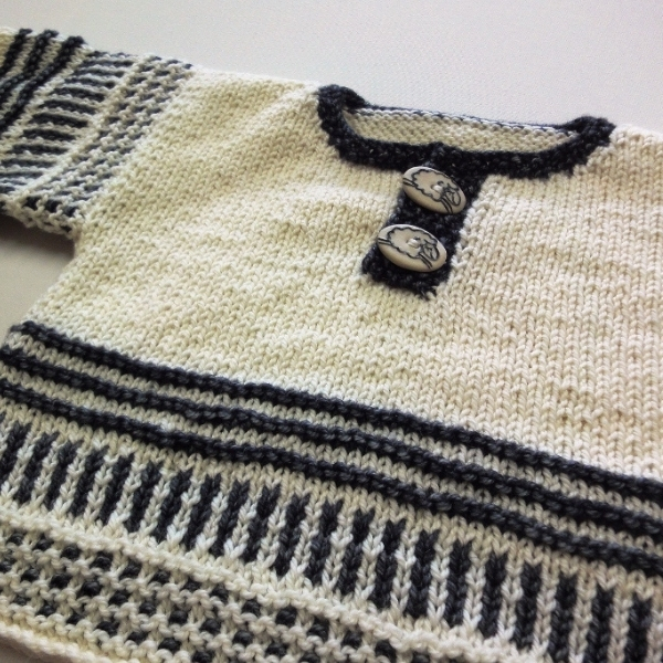 Close up of a hand knitted cream baby pullover with charcoal grey striped colourwork and two lamb motif buttons at the neckline