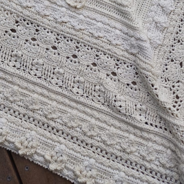 Close up of the border section of Mixology Edition 6 by Hooked on Sunshine, crocheted in Cotton Scintilla DK, draped over a wooden surface, showing alternating rows of bobble clusters, lace eyelets and textured stitch bands
