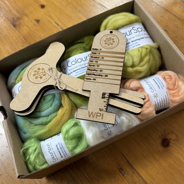 ColourSpun Tour de Fleece Mystery Box showing hand dyed Pure Merino, Kid Mohair and Tussah Silk fibre in soft green, cream and peach tones, with a flat pack Niddy Noddy and WPI measurement tool, packed in a kraft box.