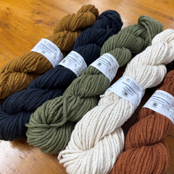 Five skeins of ColourSpun Pure Cotton Super Chunky Smooth yarn in mustard, navy, sage green, cream and rust, fanned out on a wooden table in a craft shop