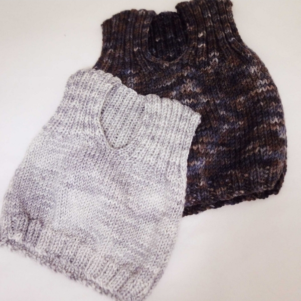 Two hand knitted children's vests made from the Child's V-neck Vest Pattern in ColourSpun hand dyed DK yarn, one in soft grey and one in dark brown tonal, laid flat on a white surface.