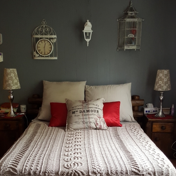 Aran Caresse Cable Afghan by Hilda Steyn spread across a bed in a grey bedroom, with red cushions and a Paris print pillow